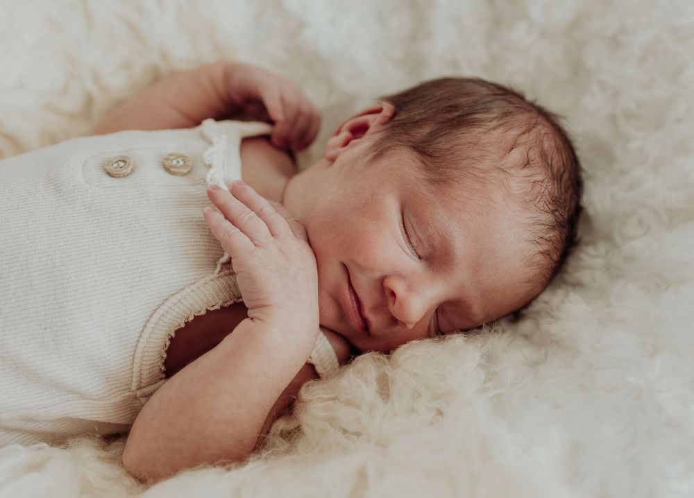 Peaceful sleeping newborn baby in cream knit romper resting on fluffy white blanket.