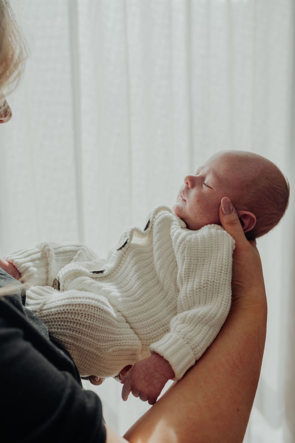 Newborn baby in a white knit outfit being cradled gently in an adult's arms indoors.