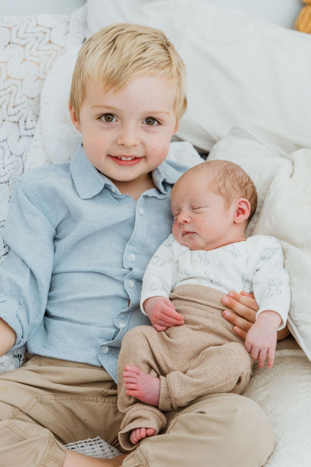 Smiling toddler boy in blue shirt holding sleeping newborn baby on white bed.