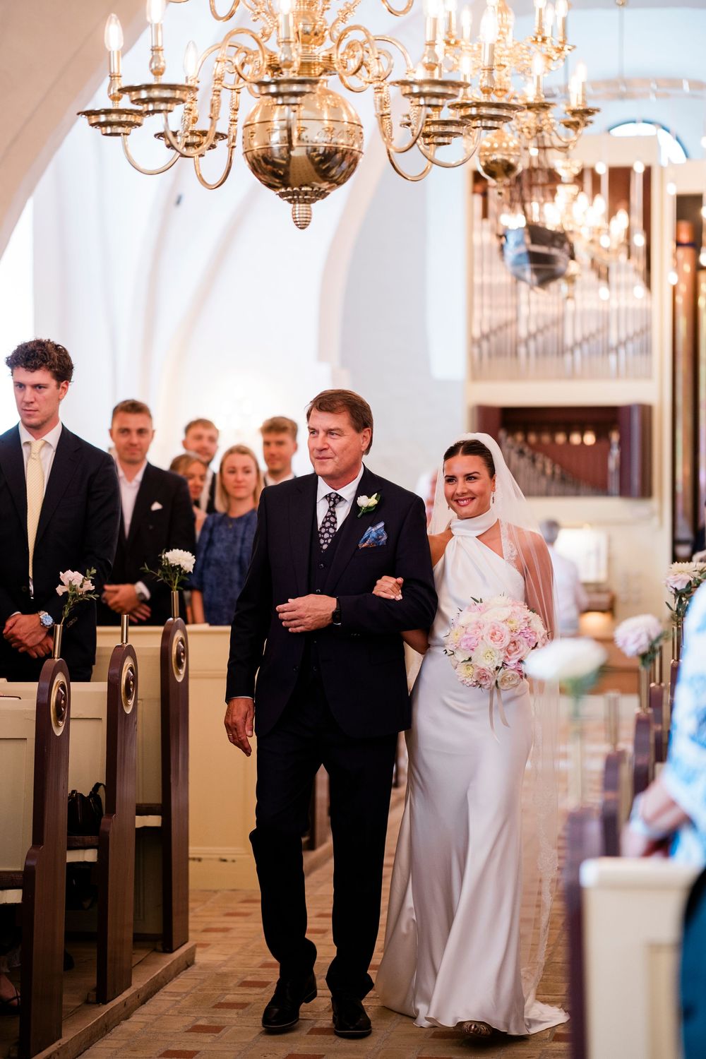 Bride in white gown walks down church aisle with father, holding bouquet, guests watching in elegant chandelier-lit venue.