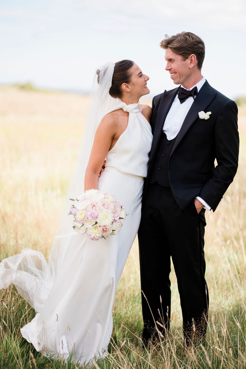Bride in white halter gown holding pink bouquet stands with groom in black tuxedo in golden field.