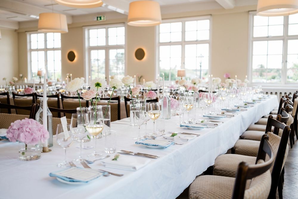 Elegant wedding reception hall with long white-clothed tables, pink floral centerpieces, crystal glasses, and blue napkins in bright venue.