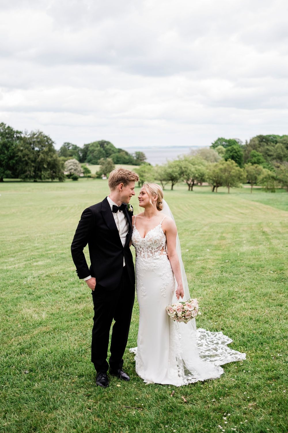 Bride and groom share a romantic moment on a lush green lawn, she in a white gown holding a bouquet, he in a black tuxedo.