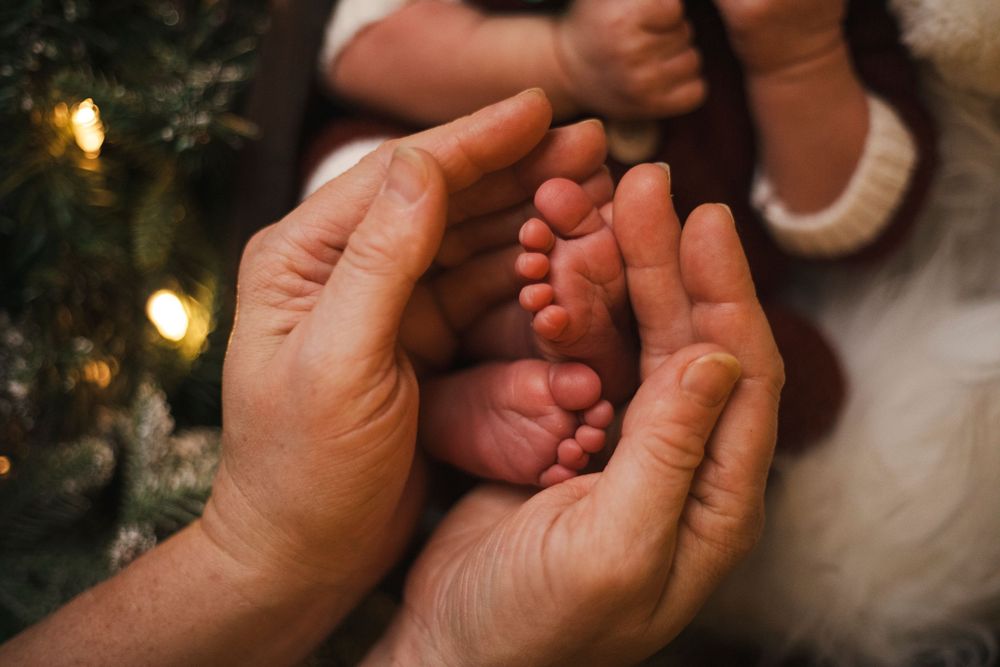 Adult hands gently cradling tiny newborn baby feet near a Christmas tree with warm glowing lights.