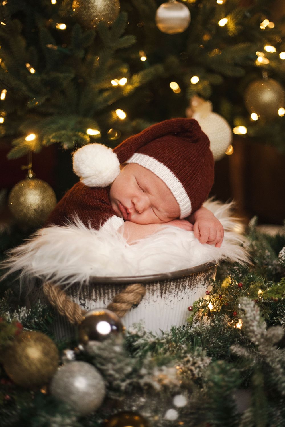 Newborn baby sleeping in a basket wearing a Santa hat surrounded by Christmas tree lights and ornaments.