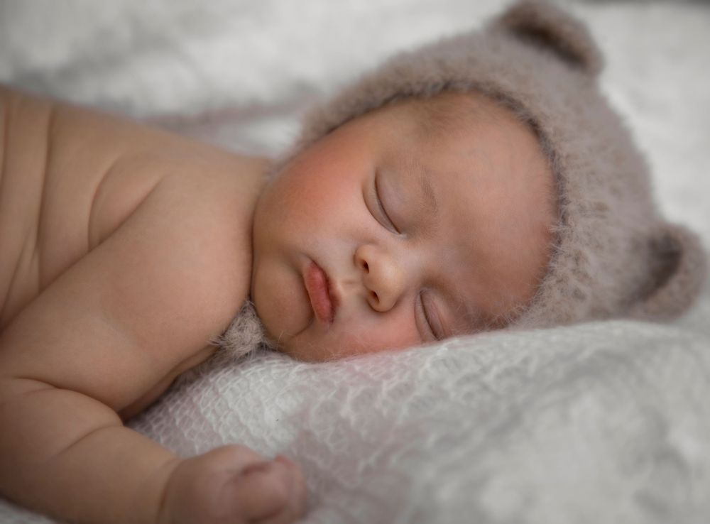 Peaceful newborn baby sleeping in a fuzzy bear ear hat on a soft white blanket.