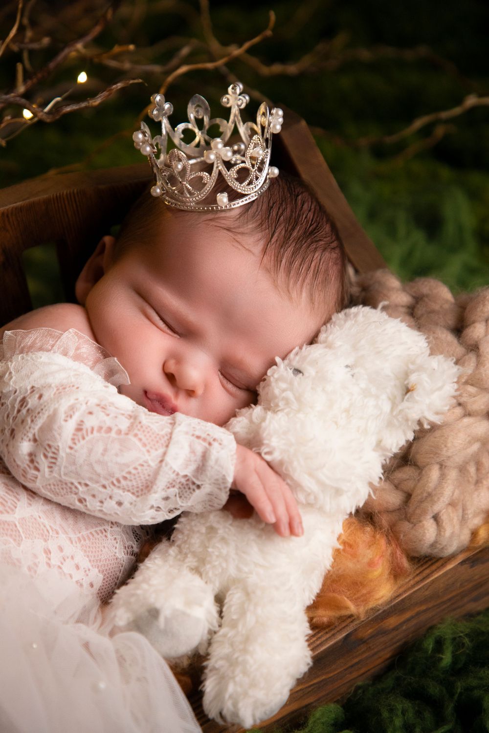 Newborn baby sleeping peacefully in white lace dress with crystal crown on wooden surface.
