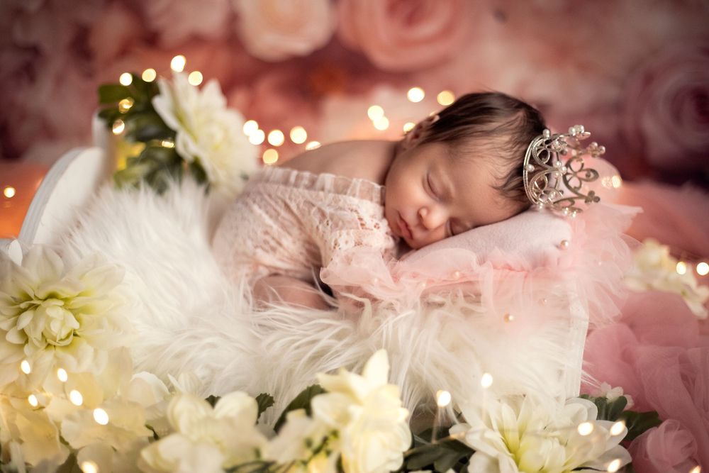 Newborn baby sleeping peacefully in a pink floral arrangement with soft bokeh lights and white flowers.