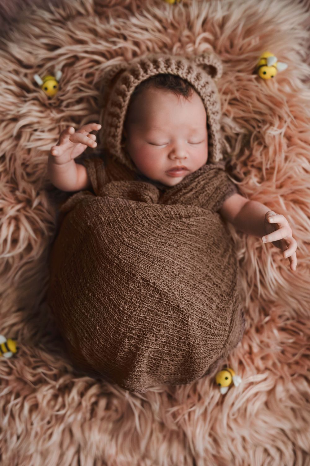 Sleeping newborn baby wrapped in brown knit swaddle wearing bear bonnet on fluffy pink fur rug with bee decorations.