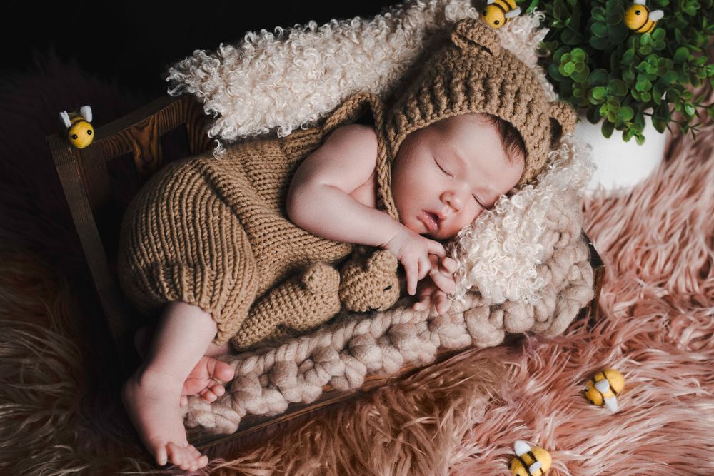Newborn baby sleeping peacefully in knit bear outfit inside a rustic wooden box with bee decorations.