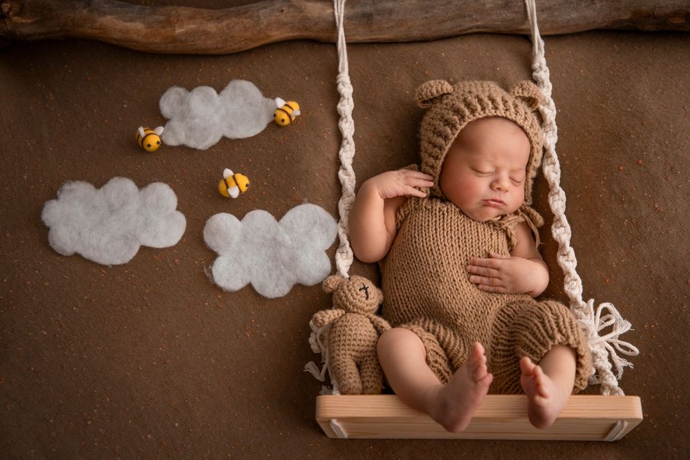 Sleeping newborn in brown knit bear outfit on wooden swing with plush clouds and bees in studio photo.