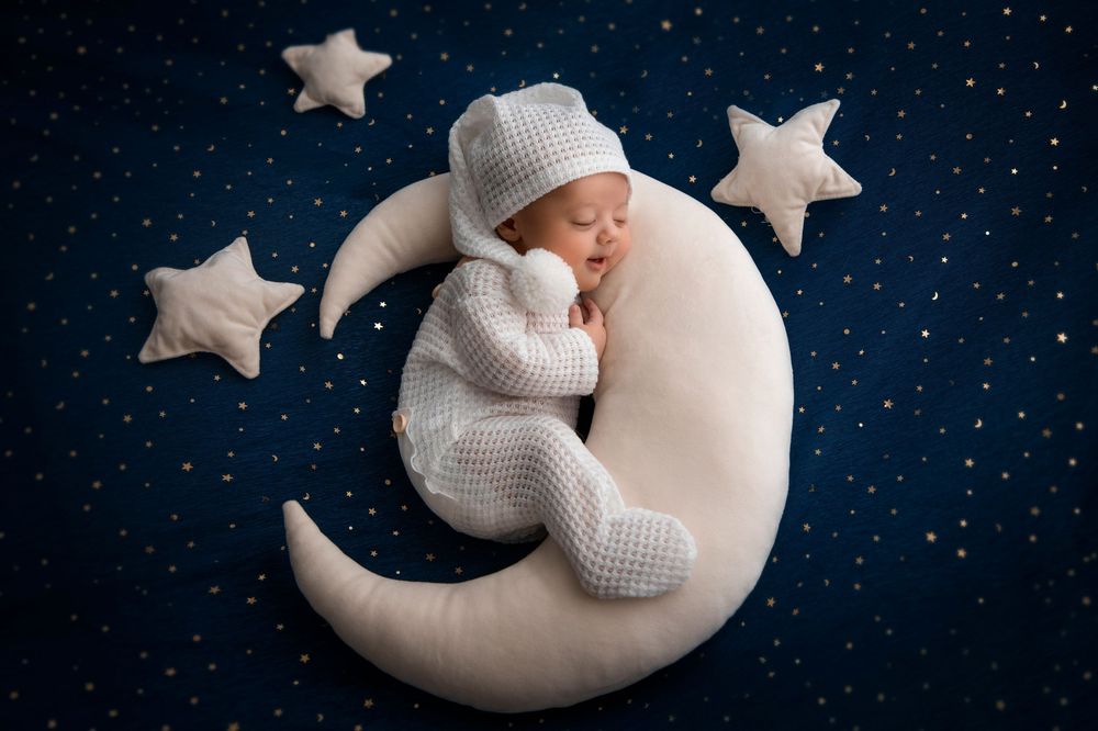 A newborn baby in white knit outfit sleeps peacefully on a crescent moon prop with stars on dark blue background.