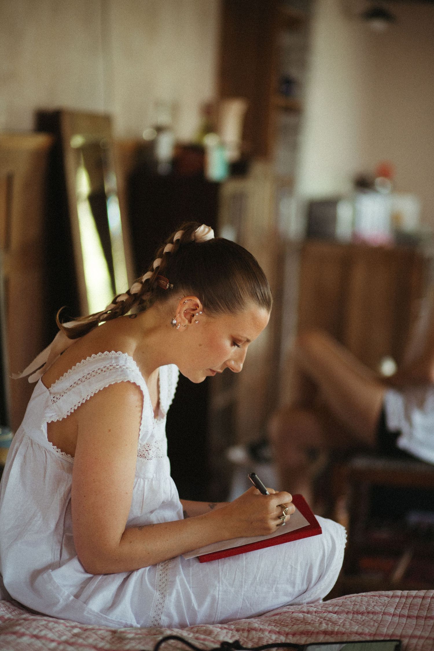 Someone in a white sleeveless dress sits on a bed writing in a red notebook with a braided hairstyle.