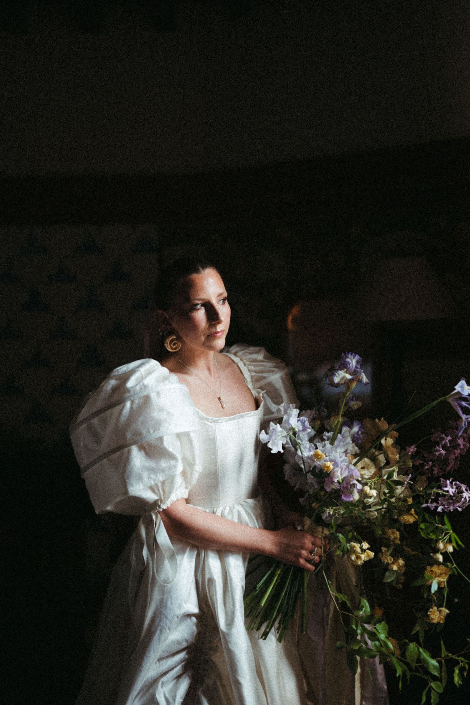 A figure in a white vintage dress holds a bouquet of colorful wildflowers against a dark background.