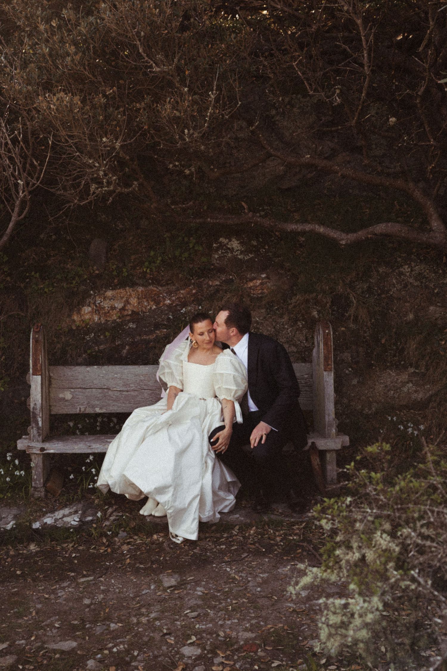 A couple in wedding attire share an intimate moment while sitting on a rustic wooden bench in a natural setting.