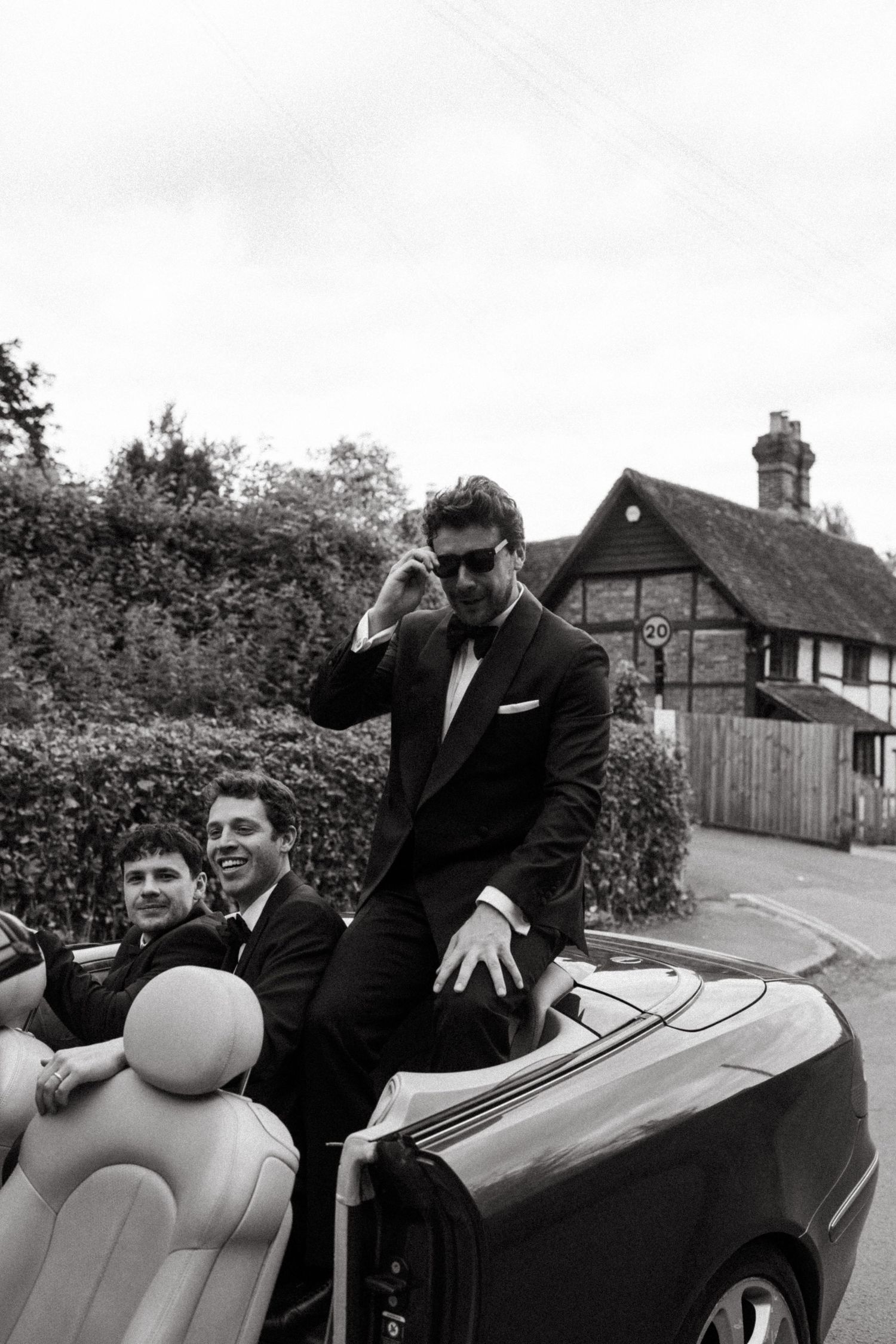 Black and white photo of people posing with a classic convertible car in front of suburban houses.