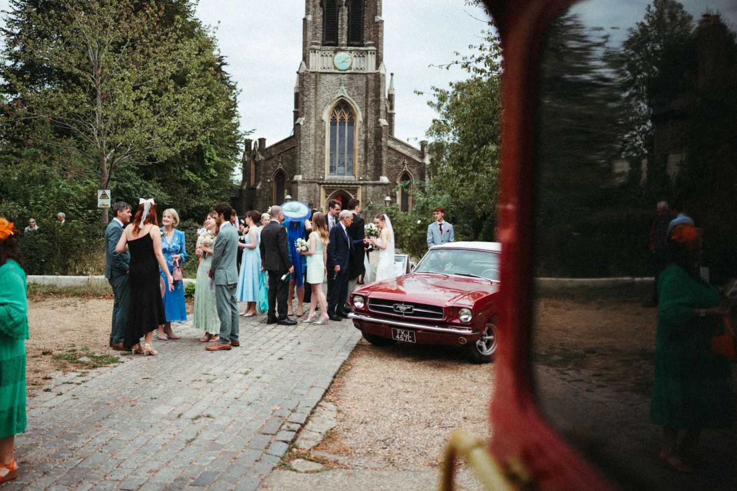 A vintage red car parked near a church with people gathering outside on a sunny day.