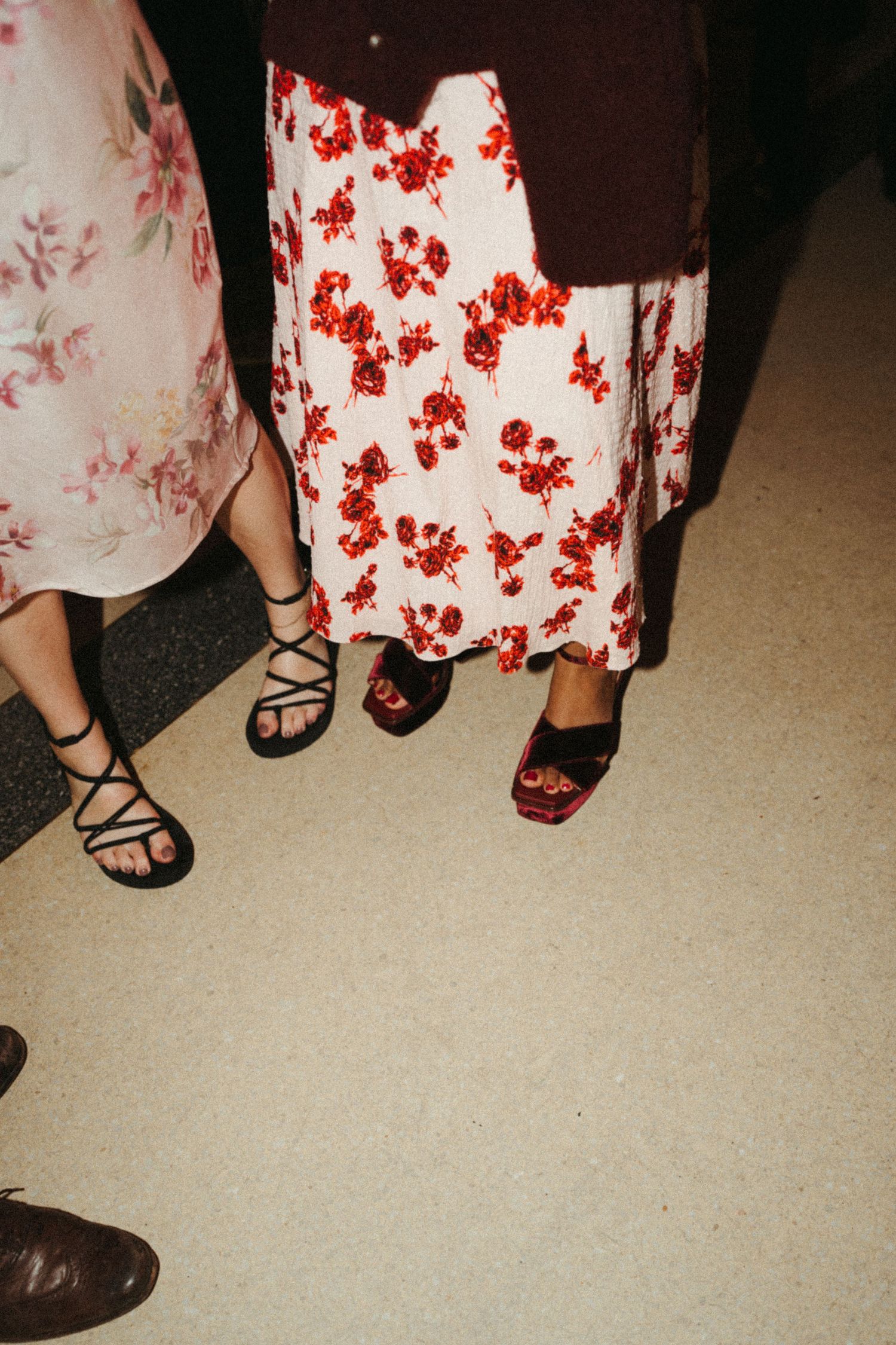 A row of women's feet wearing various sandals and shoes with a red and white floral dress pattern visible above.