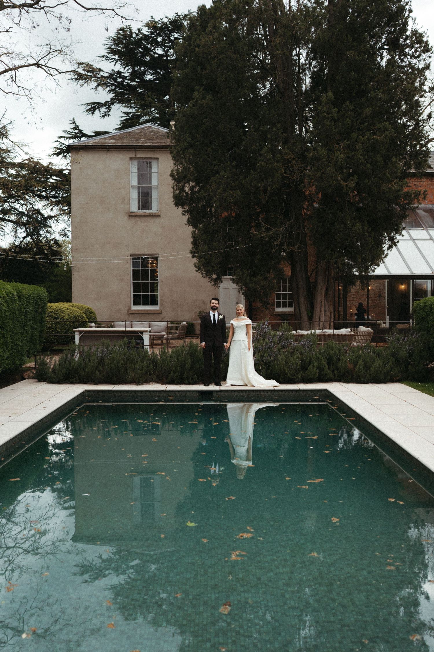 Bride and groom standing beside a reflective pool in front of an elegant estate surrounded by lush gardens.