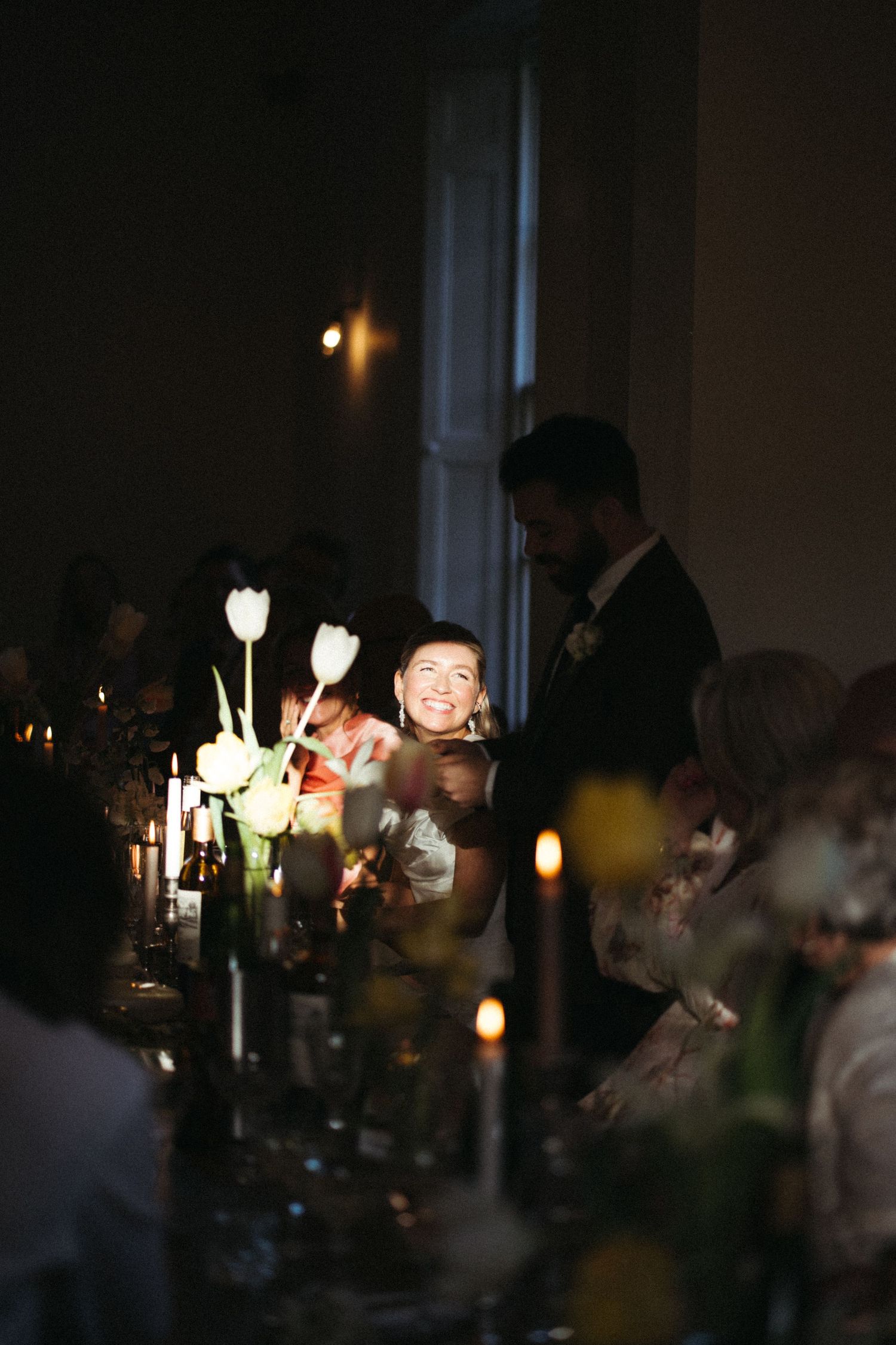 Bride smiling joyfully at candlelit wedding reception dinner table with tulips and guests in dim romantic setting.