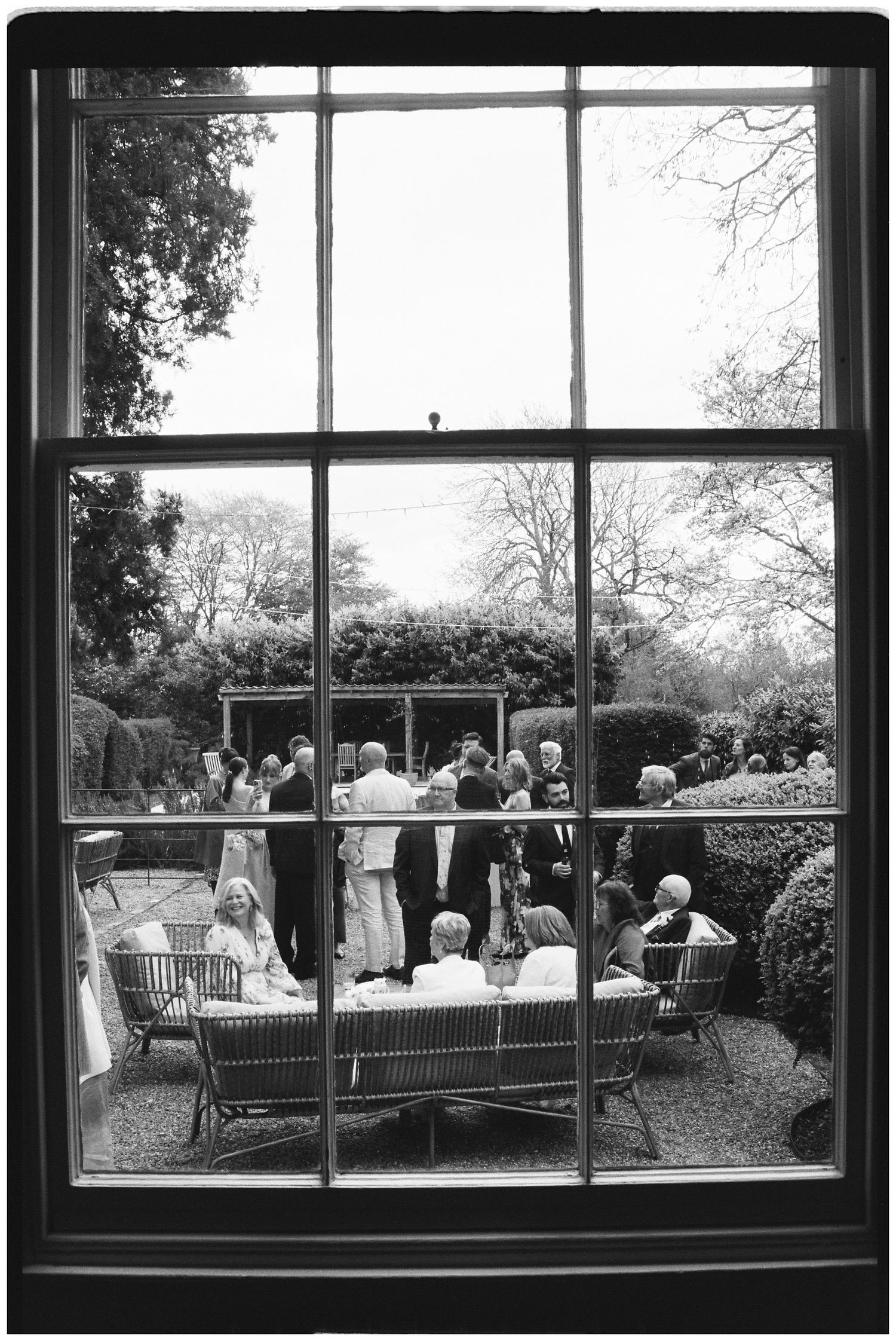 Black and white photo of garden party guests socializing on a patio, viewed through a multi-pane window.