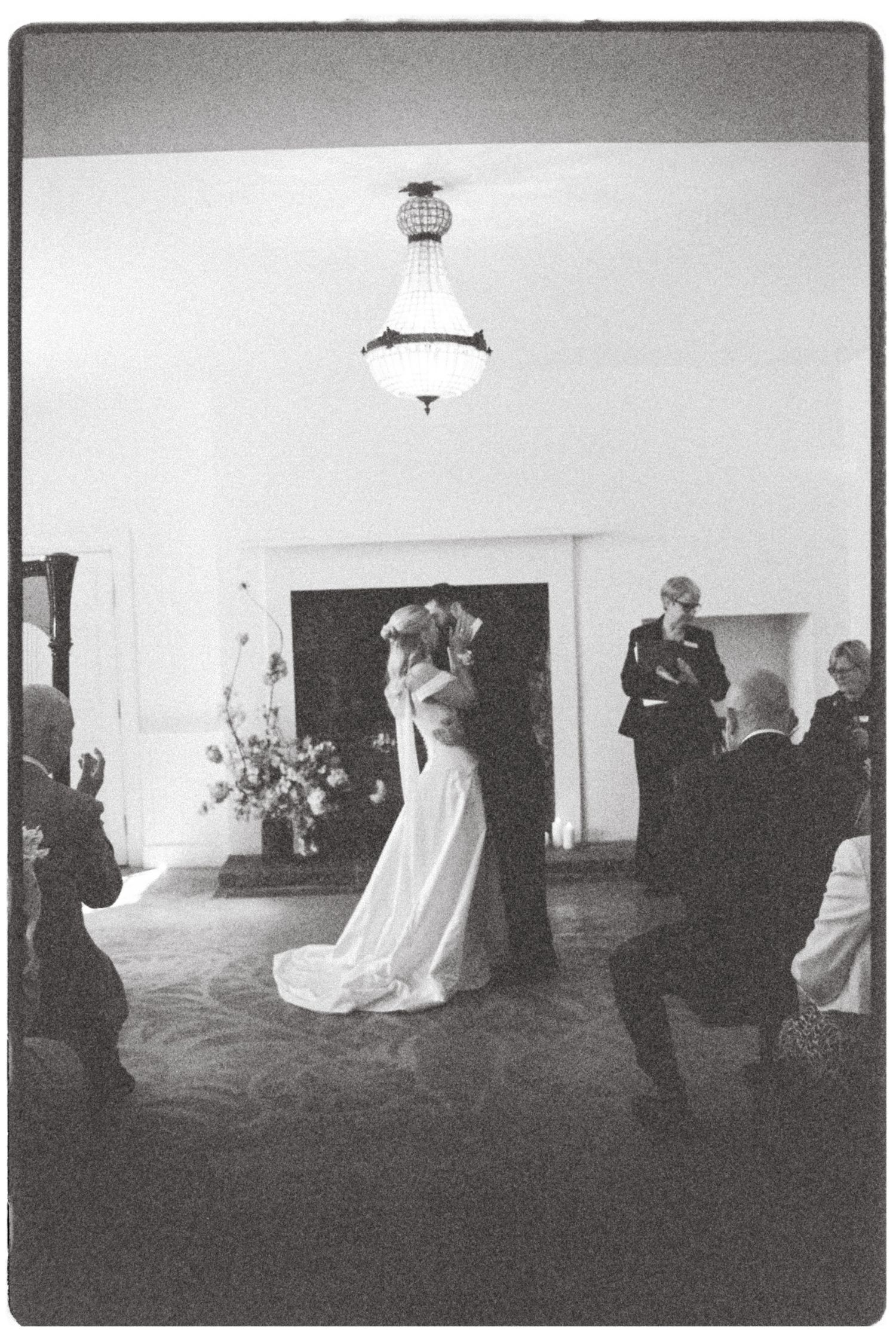 Black and white wedding photo of bride and groom embracing during ceremony with guests and floral arrangement in background.