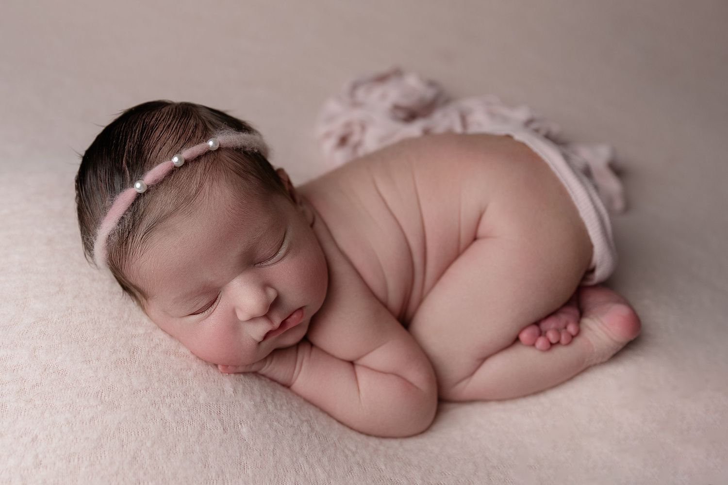 Sleeping newborn baby in delicate lace headband curled up on soft pink blanket during peaceful professional photoshoot.