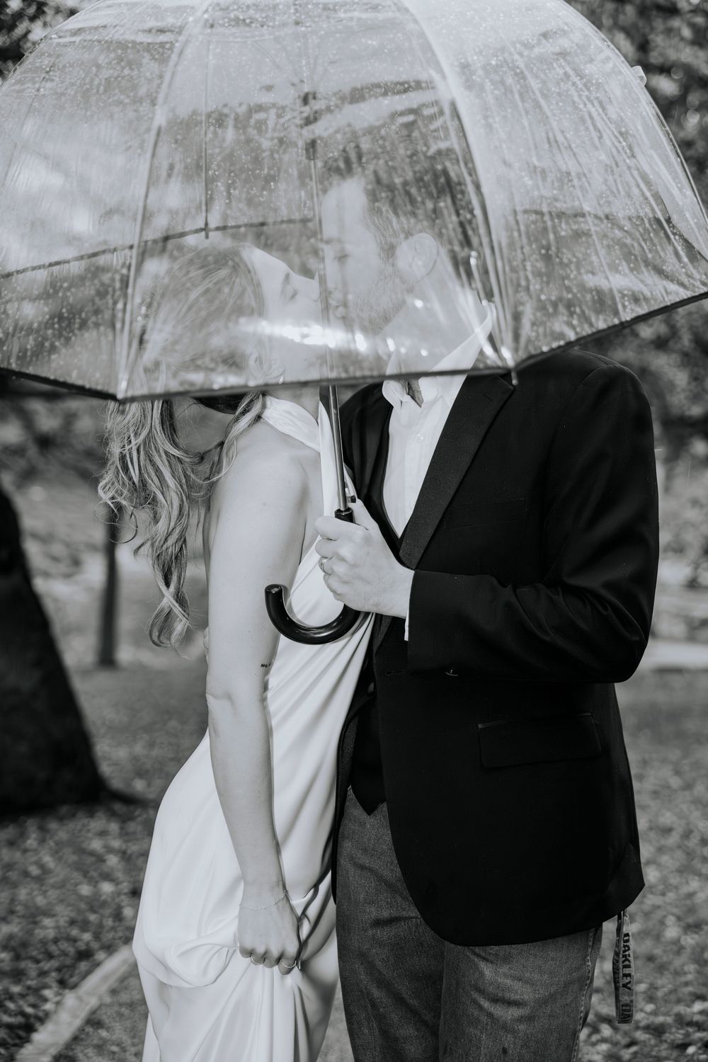 Black and white photo of bride and groom sharing a clear umbrella in the rain on their wedding day.