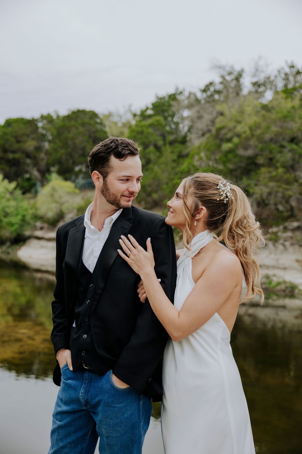 A bride in a white halter dress and groom in a black blazer share a romantic moment by a serene river.