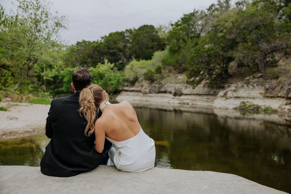 Bride and groom sitting together on a rock by a serene river, surrounded by lush green trees.
