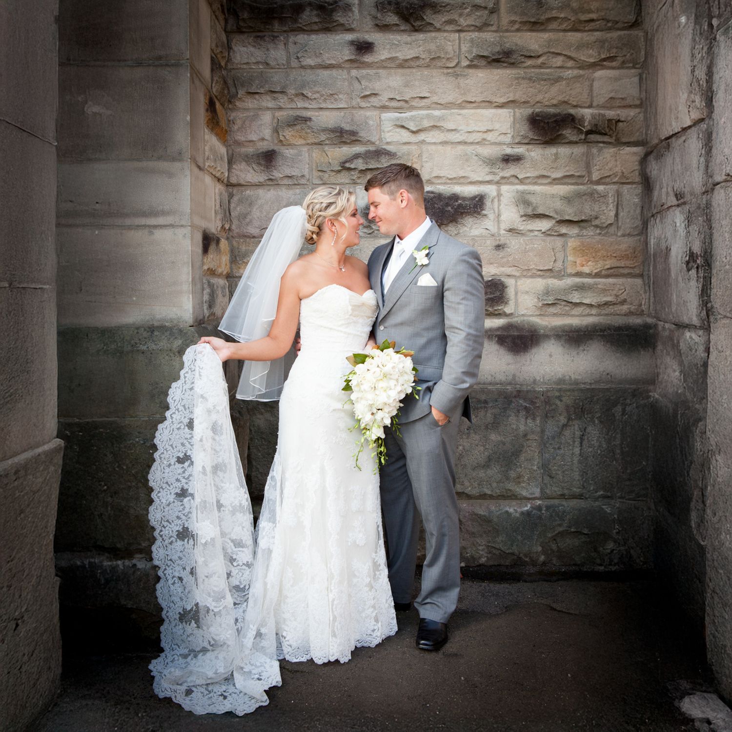 Bride holding long lace trim veil standing with groom in grey suit by stone wall, East Anglia wedding photographer.