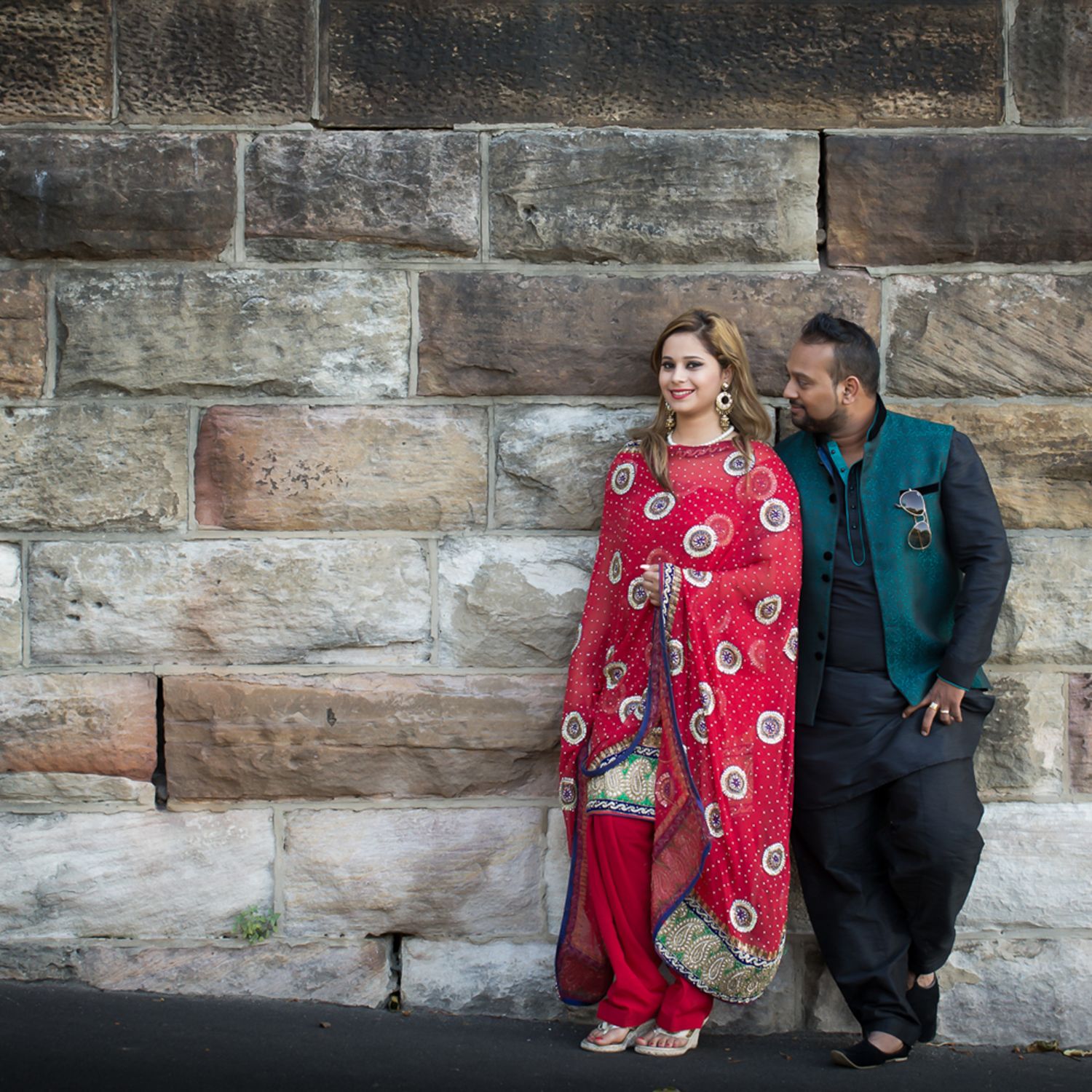 Indian bride and groom in traditional red and teal outfit leaning against sandstone wall, London wedding photography.