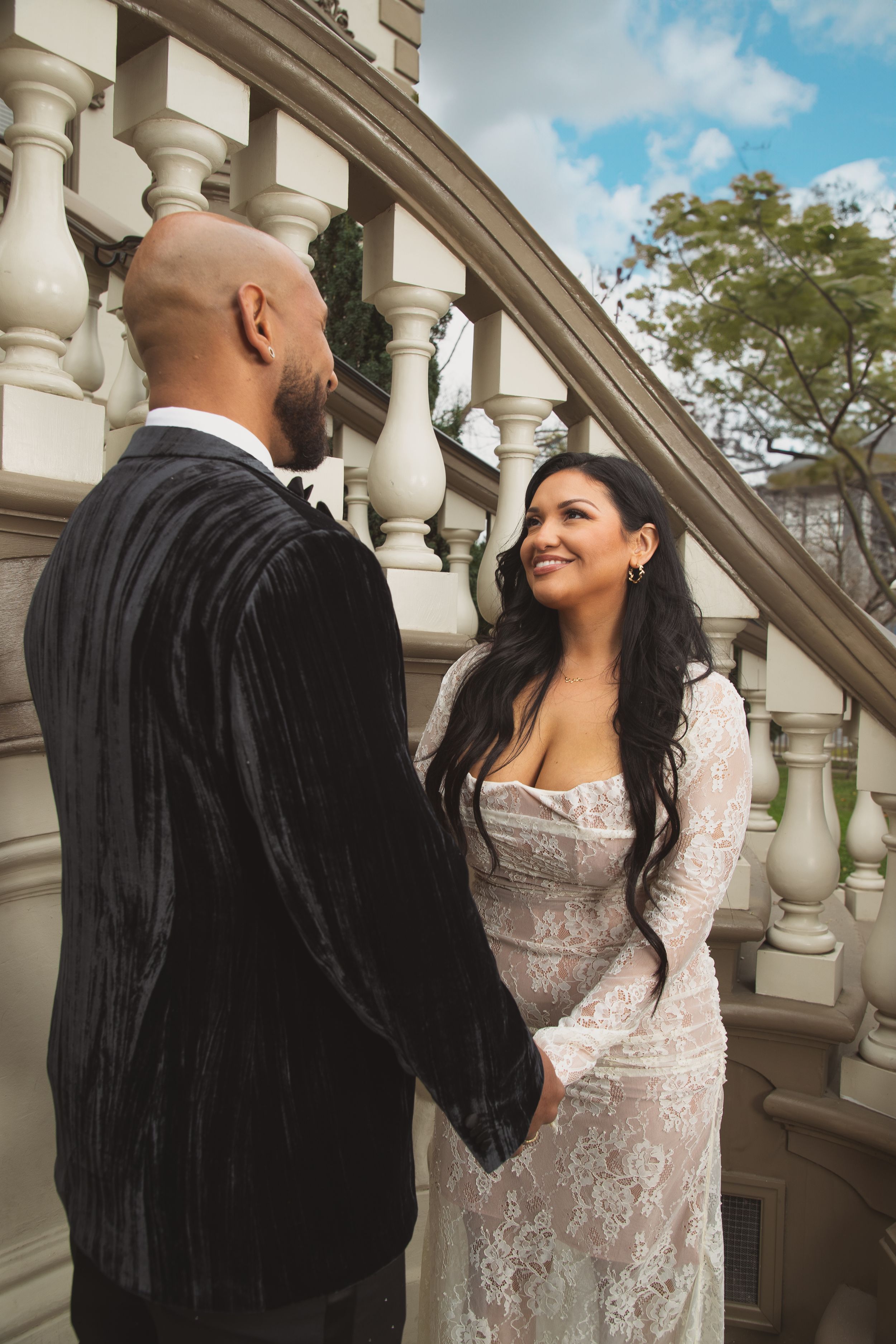 Couple in formal attire smile at each other while standing on elegant stone staircase with ornate balustrades.