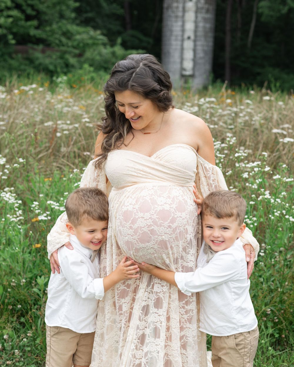 A maternity photo in a wildflower field shows two young boys in white shirts hugging a pregnant belly.