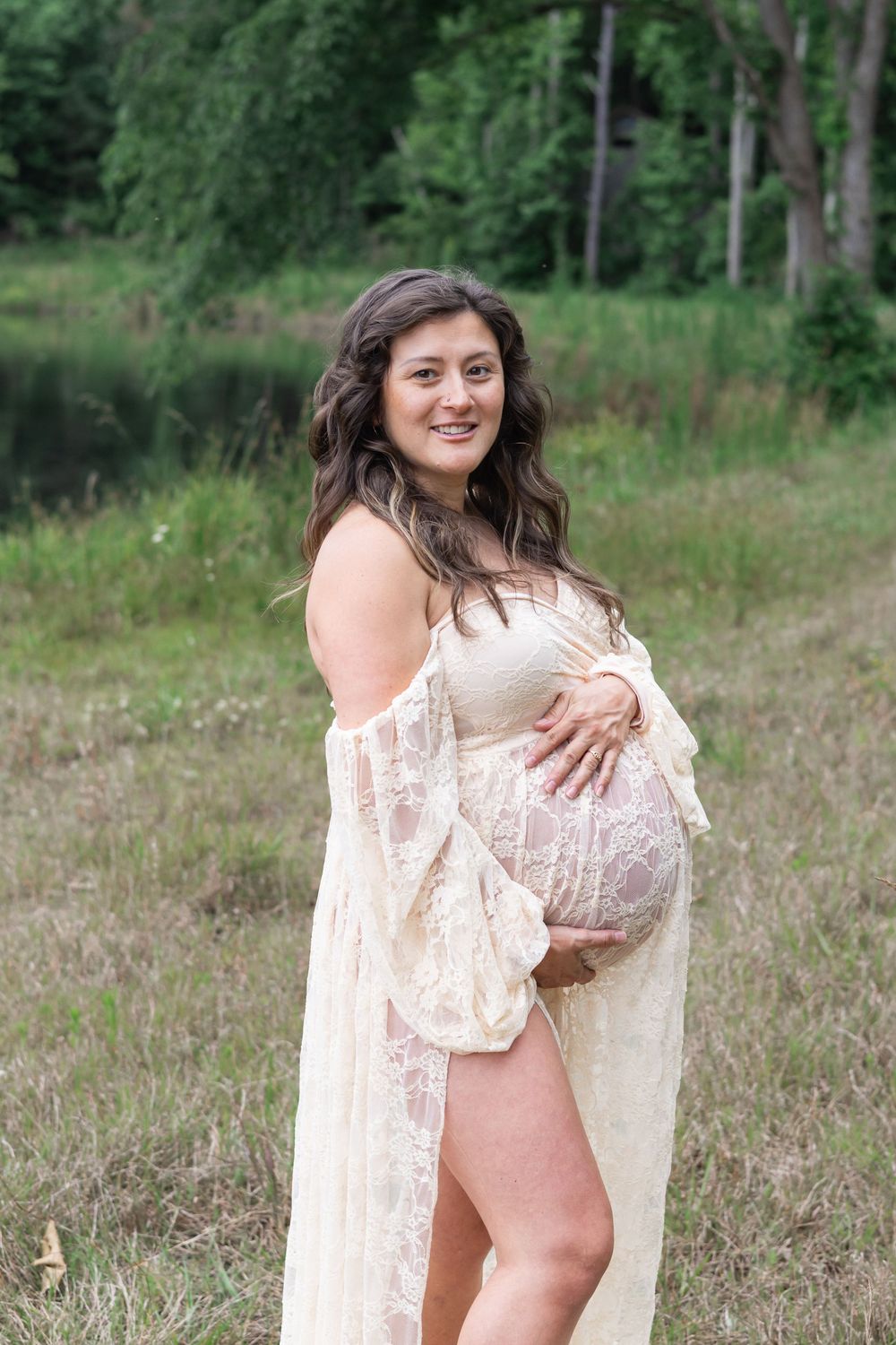 A maternity portrait in a lace dress against a natural outdoor setting with trees and grass.