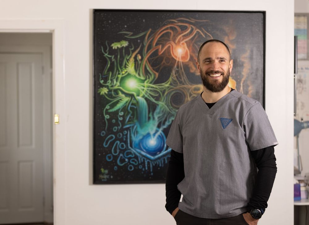 Smiling man in gray scrubs stands beside colorful cosmic artwork in a medical office setting.