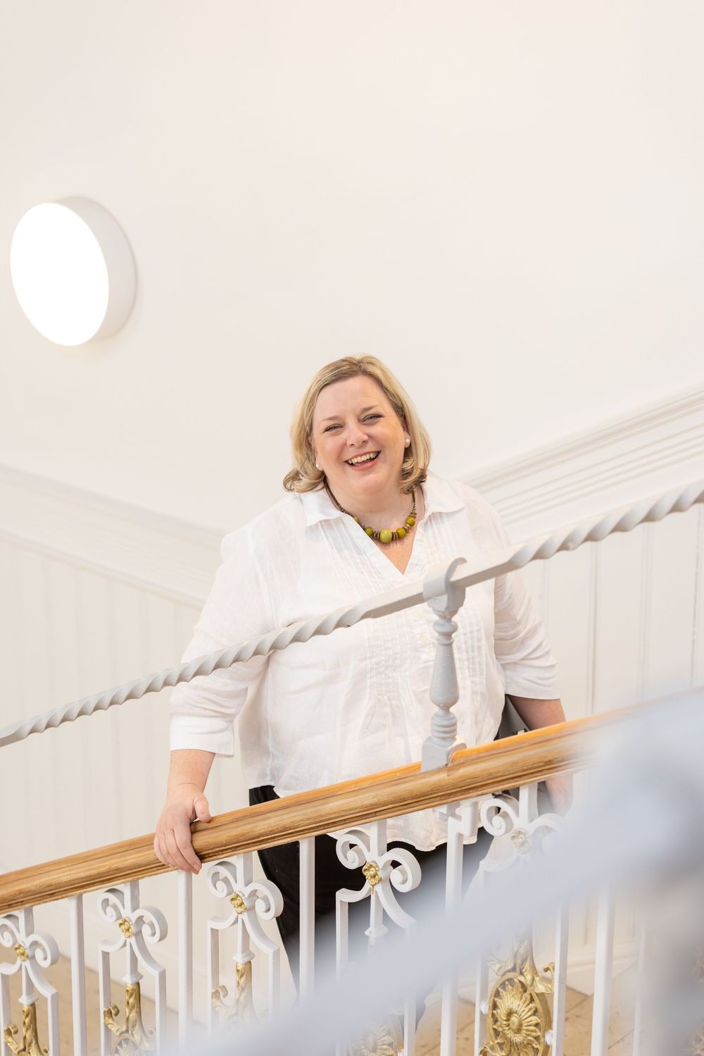 Smiling blonde woman in white blouse leaning on ornate staircase railing in elegant white interior.