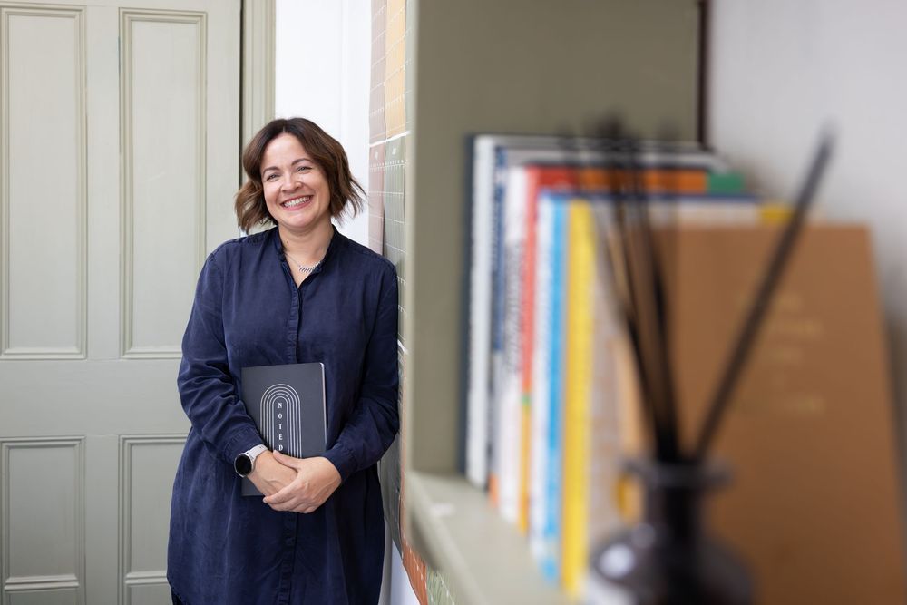 A smiling woman in a navy dress holds a portfolio while standing near a bookshelf filled with colorful books.