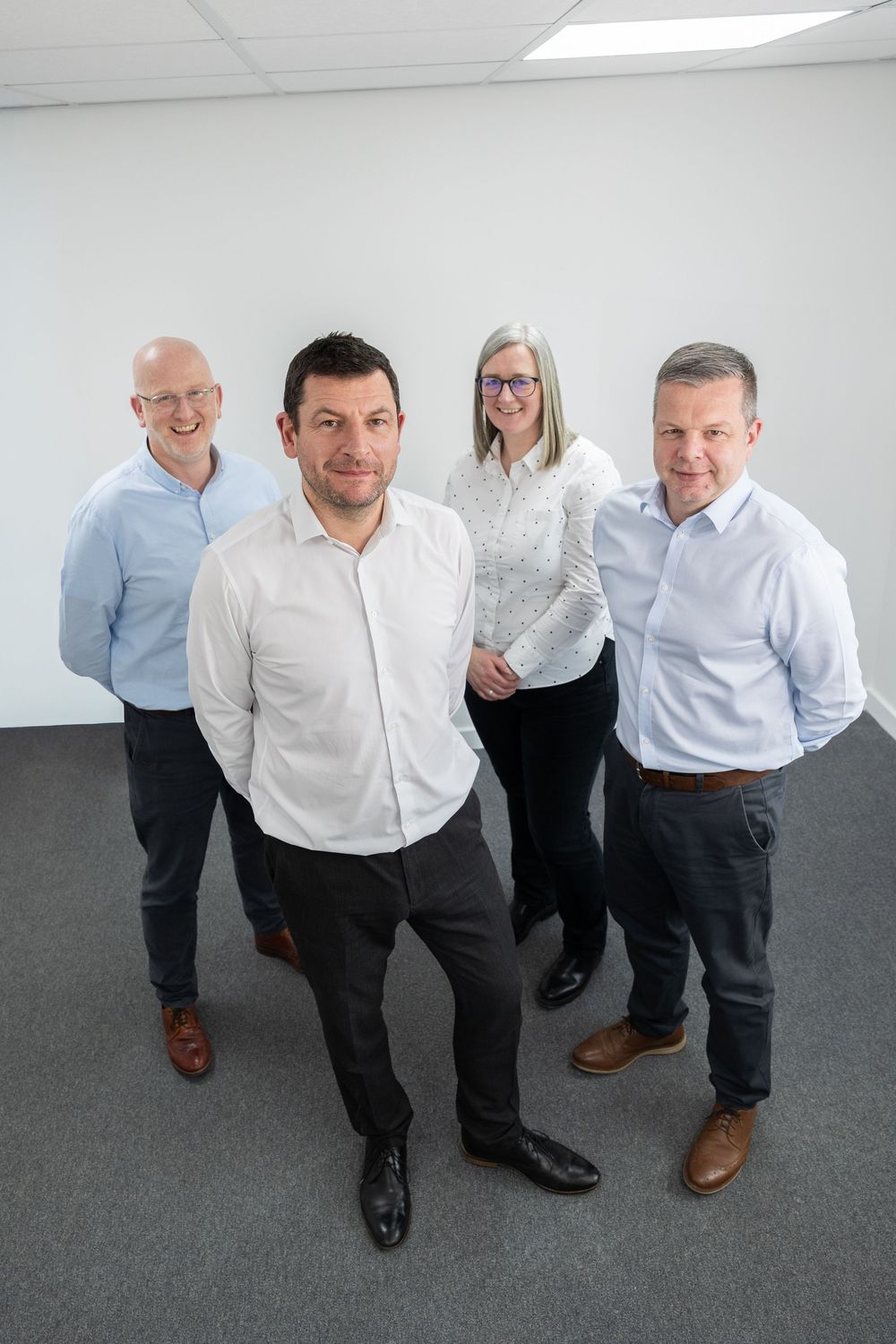 Four business professionals in smart casual attire pose together smiling in a modern office space.