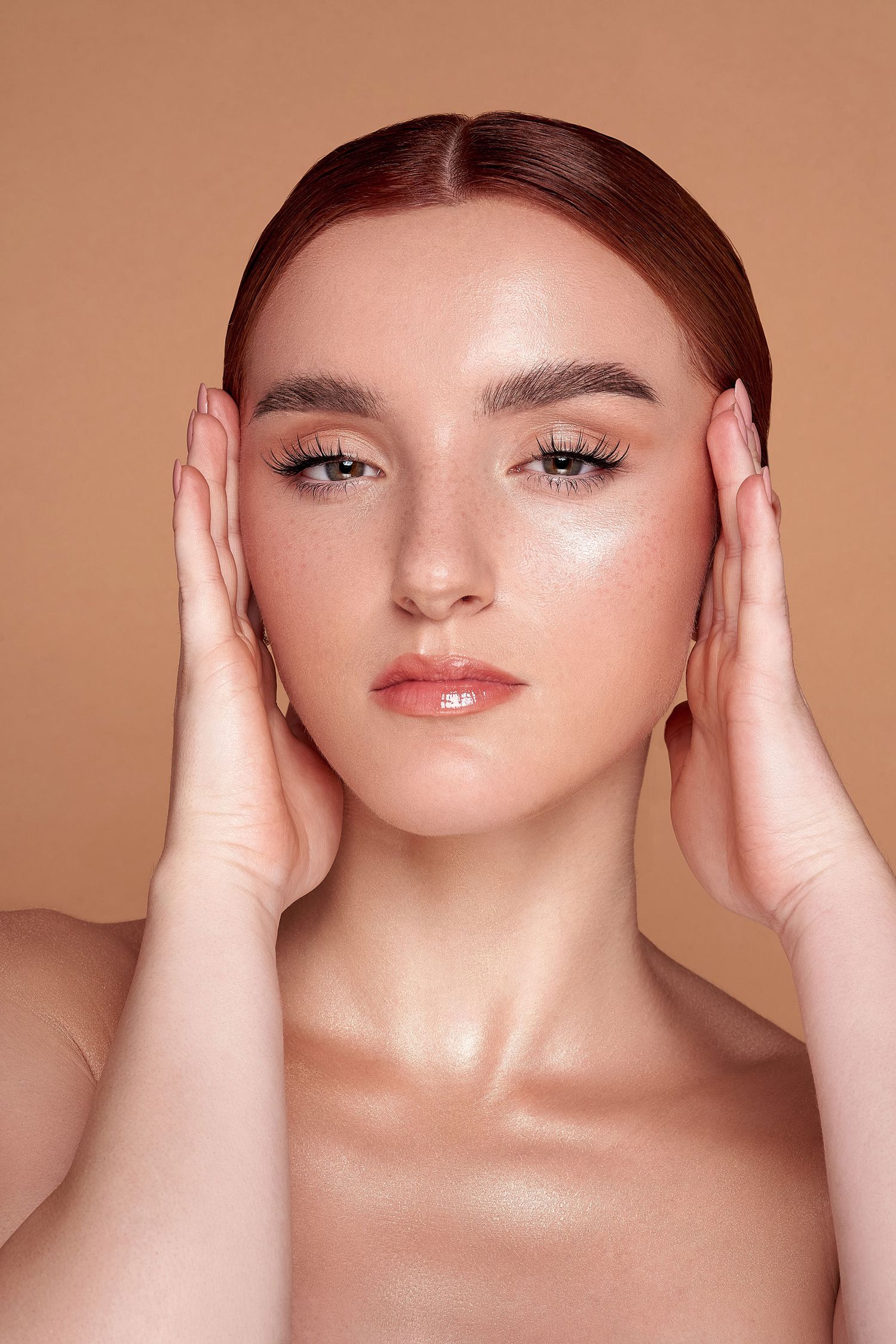 Beauty portrait with glowing makeup, slicked back hair, and hands framing face against peachy backdrop.