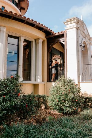 Couple embracing on a elegant Mediterranean-style home's porch surrounded by lush green shrubs and red flowers.
