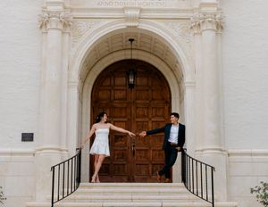Couple holding hands on steps of elegant white building with ornate arch and wooden doors.