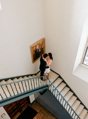 Couple sharing a romantic kiss on elegant staircase beside a framed portrait in a classic white-walled venue.