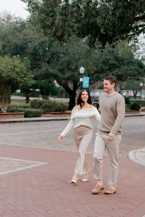 Couple walks hand-in-hand smiling in beige outfits at an outdoor brick plaza.