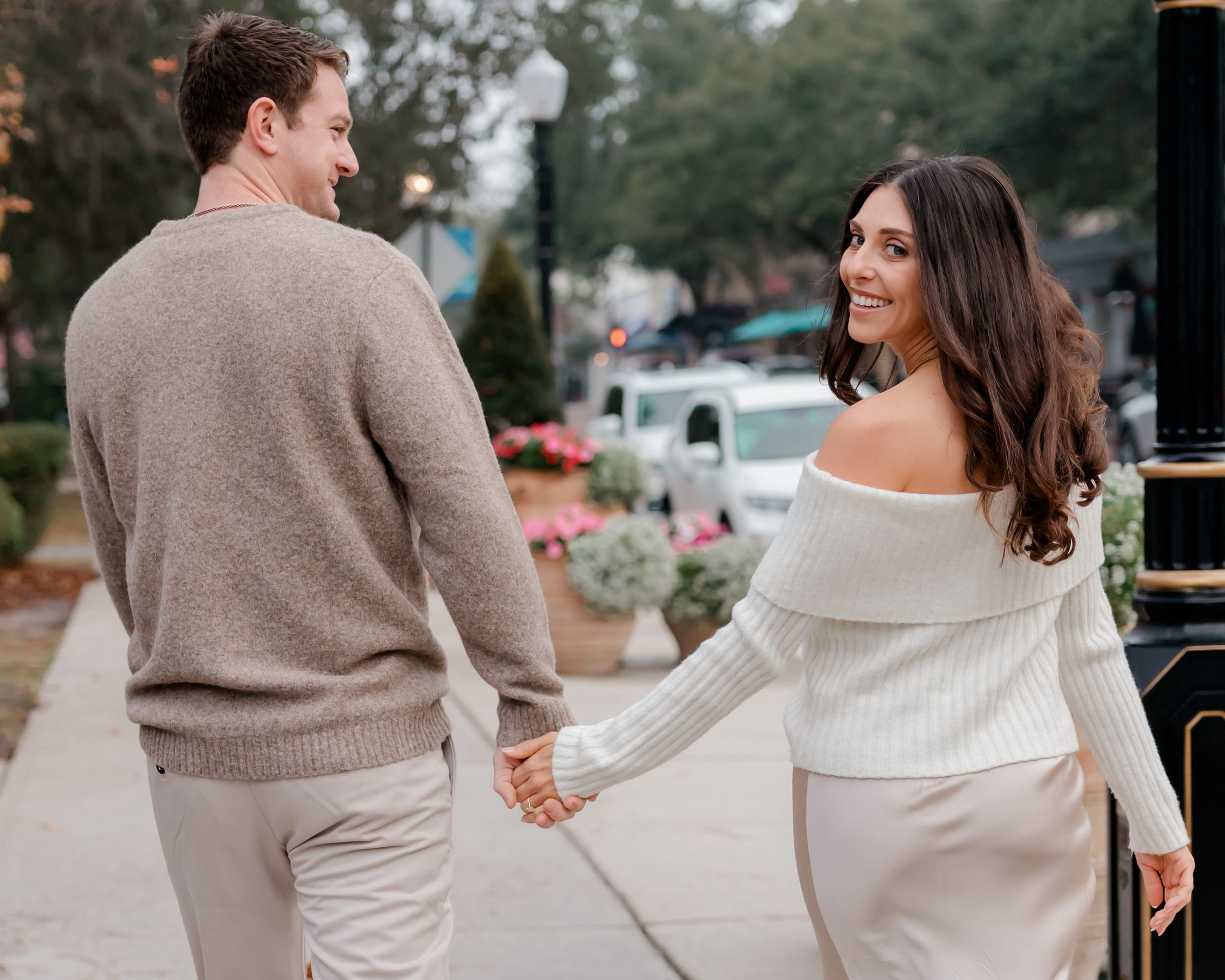 Couple holding hands on a tree-lined sidewalk, woman in white off-shoulder top smiling back at camera.
