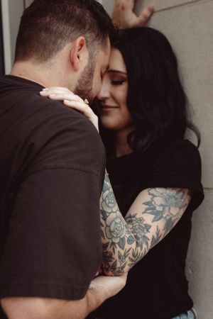 A romantic embrace shows detailed floral tattoo sleeve artwork against a black top and wall background.