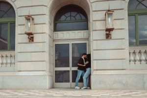 Someone leans against a glass door entrance of an elegant stone building with arched windows and decorative sconces.