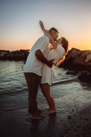 A romantic couple embraces on a beach at sunset with rocky coastline in the background.