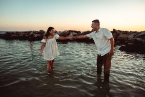 Two people wearing white clothes stand in shallow ocean water at sunset near rocky shoreline.