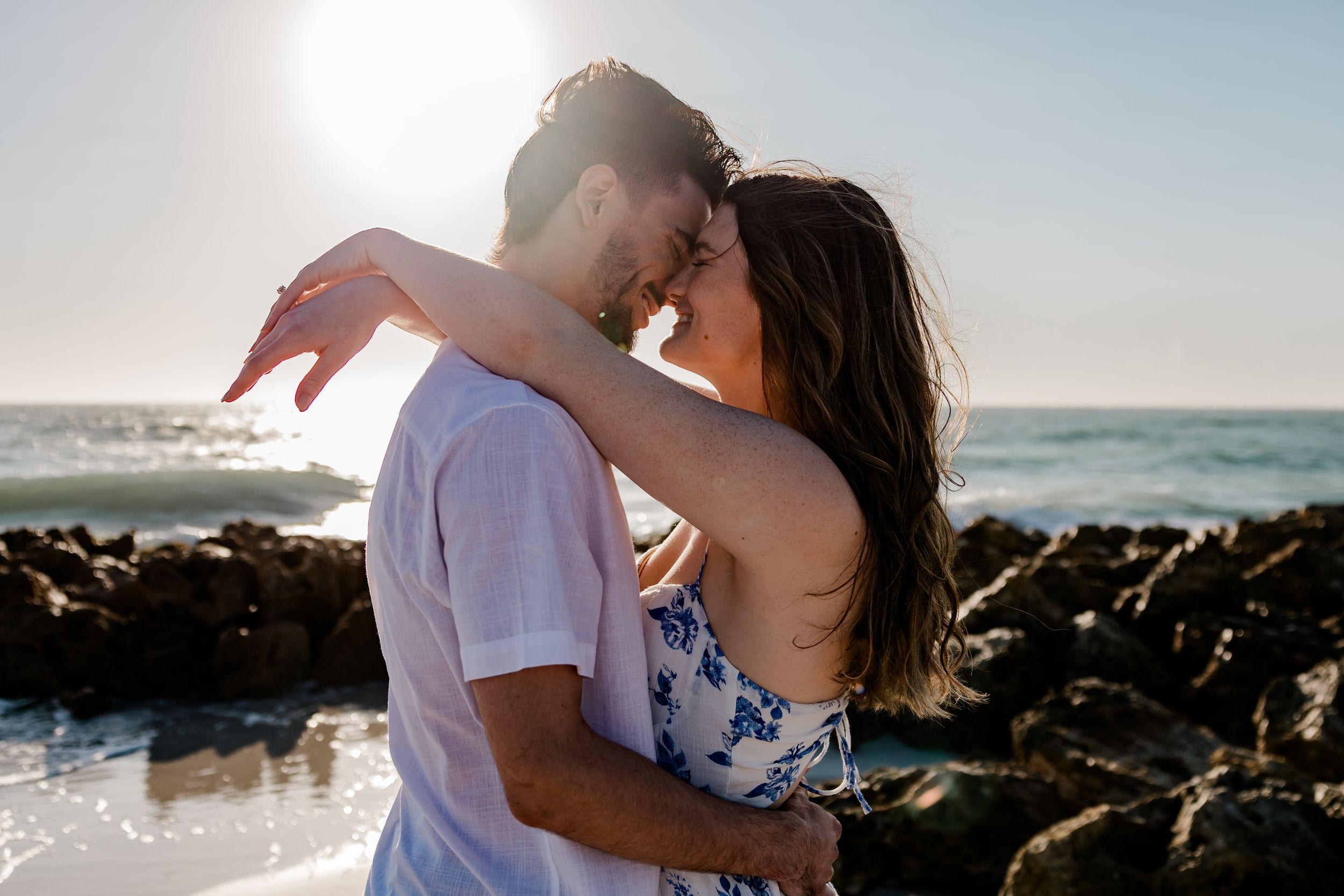 A couple embraces on a rocky beach at sunset with ocean waves and sunlight creating a romantic silhouette.